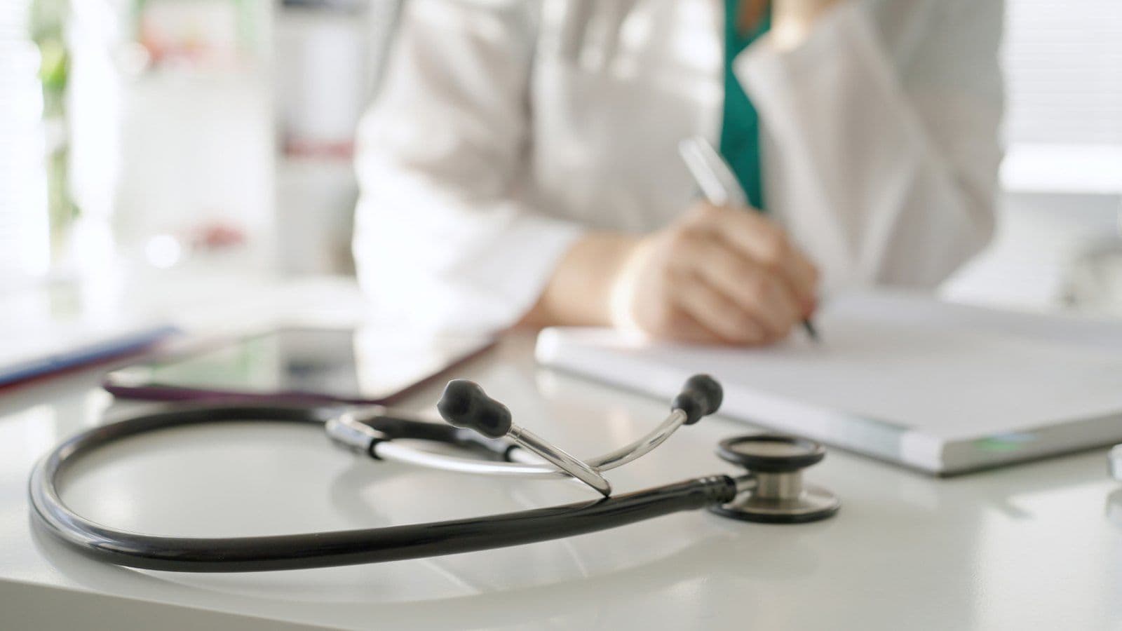 Stethoscope pose sur un bureau, medecin prenant des notes en arriere-plan