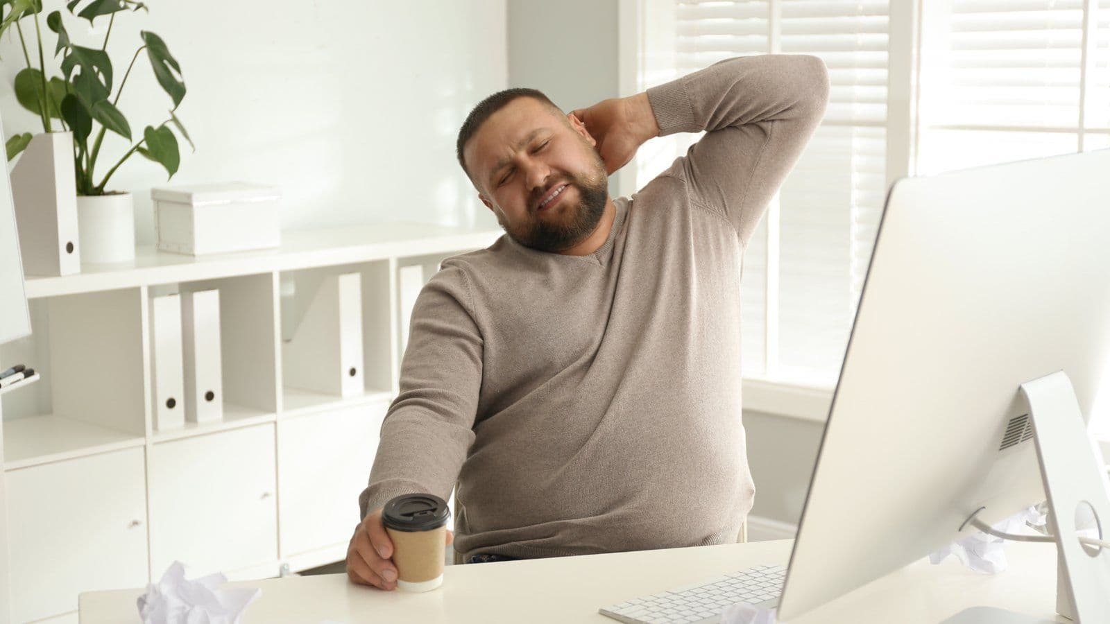 Homme se massant la nuque devant son ecran, gobelet de cafe en main