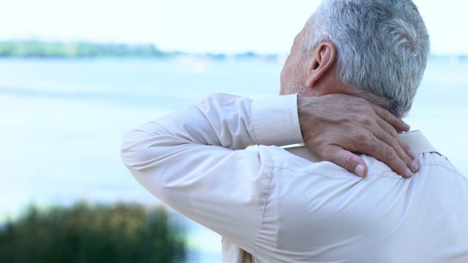 Homme senior de dos massant sa nuque face a un plan d'eau